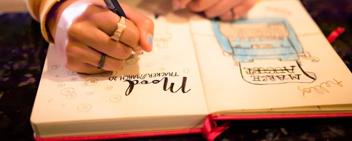 Close-up van een hand die in een bullet journal schrijft; open notitieboek met geïllustreerde lijstjes en doodles, pen en potlood op tafel.