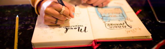 Close-up van een hand die in een bullet journal schrijft; open notitieboek met geïllustreerde lijstjes en doodles, pen en potlood op tafel.