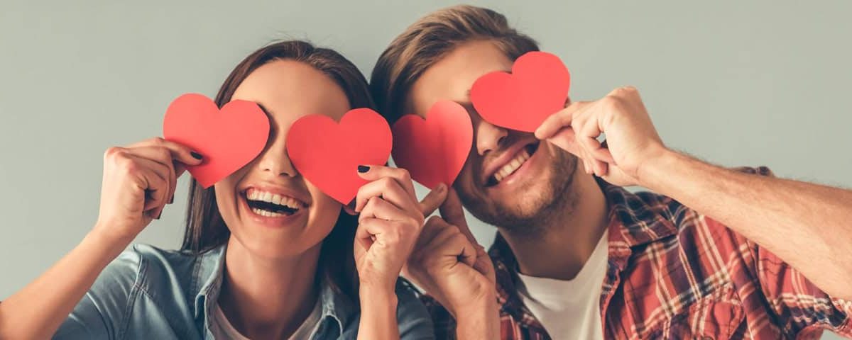 Lachend stel houdt rode papieren hartjes voor de ogen — vrolijke Valentijnssfeer.