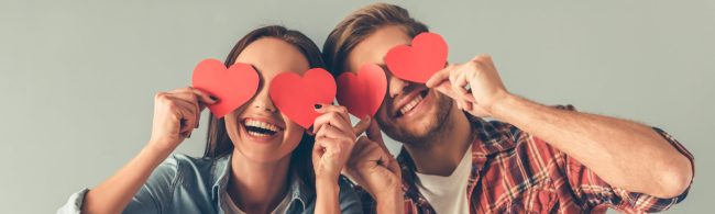 Lachend stel houdt rode papieren hartjes voor de ogen — vrolijke Valentijnssfeer.