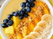 Tropical smoothie bowl met kleurrijke lagen: yoghurt met mango, perzik, blauwe bessen, ananas, pistachenoten, lijnzaad en kokossnippers.