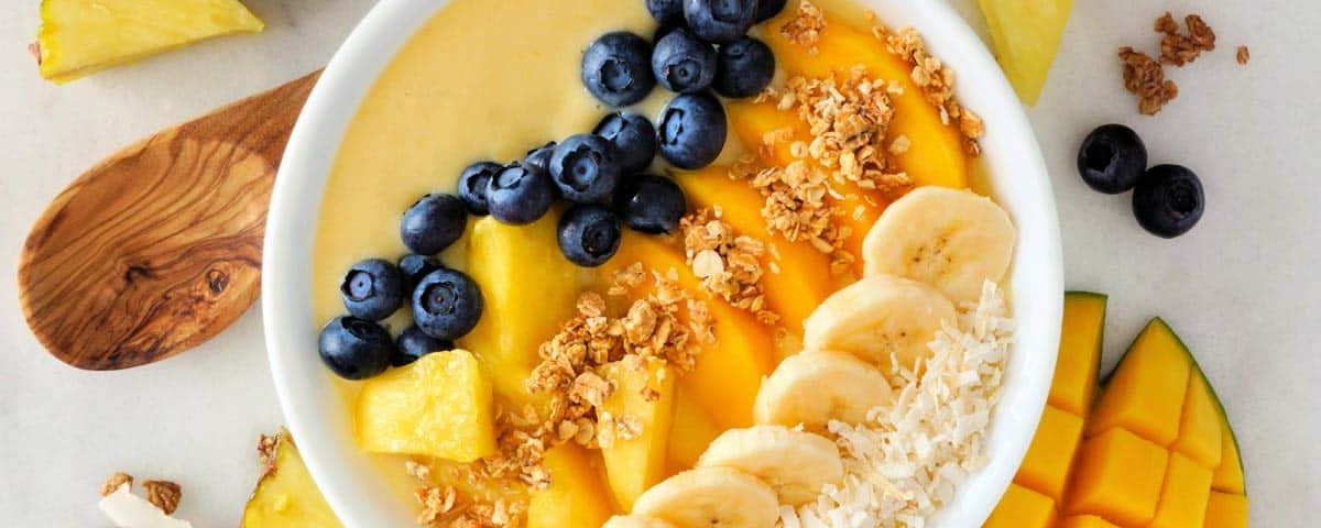 Tropical smoothie bowl met kleurrijke lagen: yoghurt met mango, perzik, blauwe bessen, ananas, pistachenoten, lijnzaad en kokossnippers.