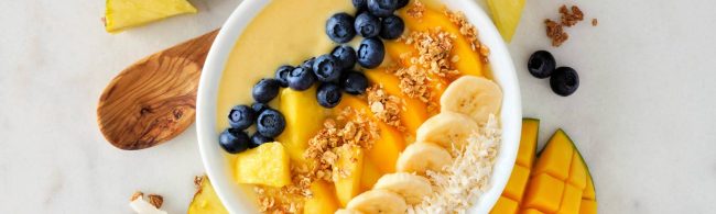 Tropical smoothie bowl met kleurrijke lagen: yoghurt met mango, perzik, blauwe bessen, ananas, pistachenoten, lijnzaad en kokossnippers.